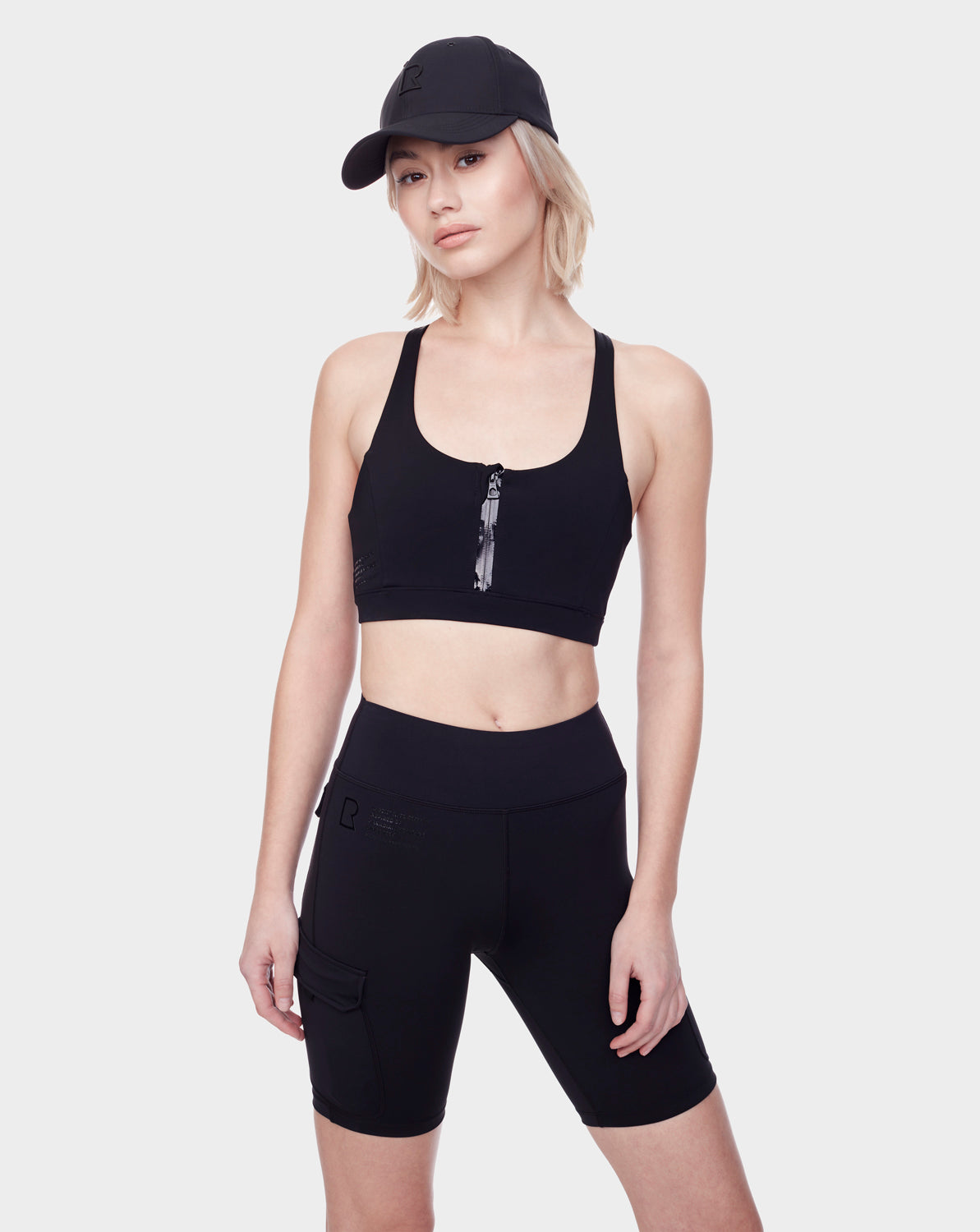 A woman wearing the Rudsak SERINA - 8521006 BLACK high-performance black sports bra and high-waisted biker shorts, paired with a black cap, stands against a plain white background, looking at the camera with a neutral expression.