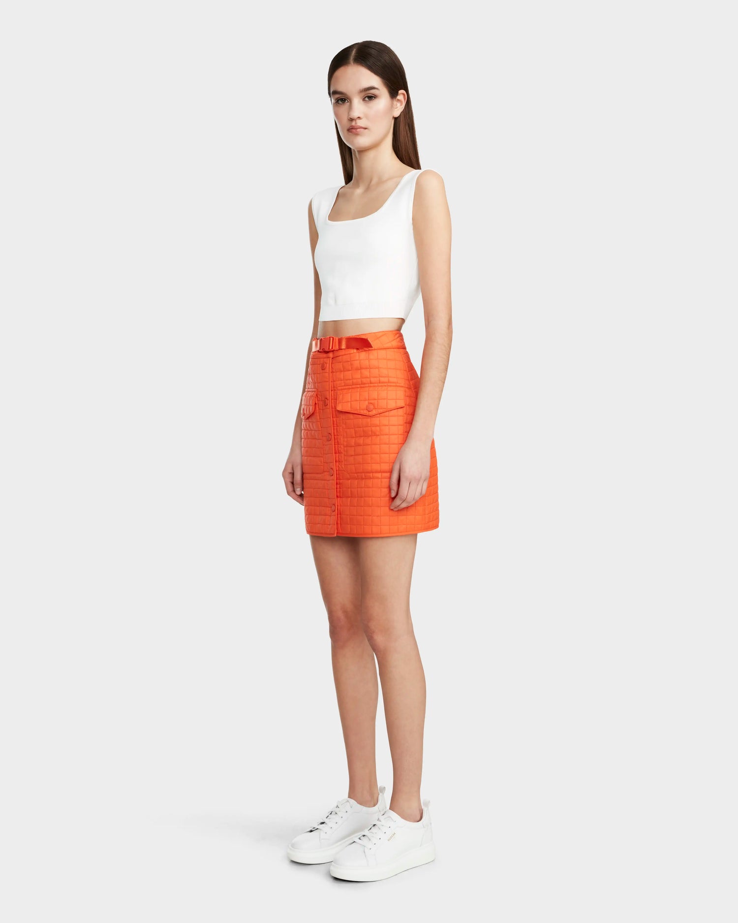 A woman stands slightly to the side wearing Rudsak's SABINE - 8524063 Orange Sorbet mini skirt with front pockets and a belt, a white sleeveless crop top, and white sneakers, set against a plain light gray background.