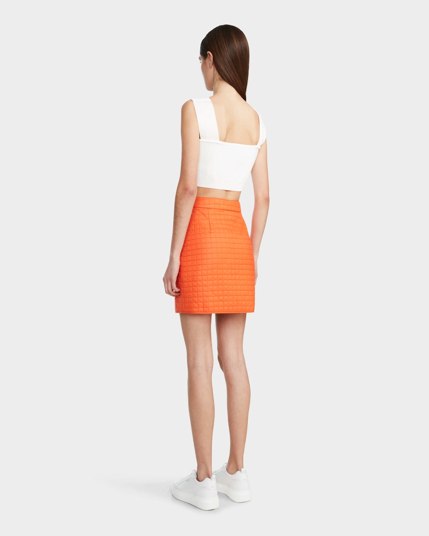 A woman with loose straight brown hair faces away, wearing a white sleeveless crop top, white sneakers, and the Rudsak SABINE - 8524063 Orange Sorbet quilted short skirt, posing against a light gray background.