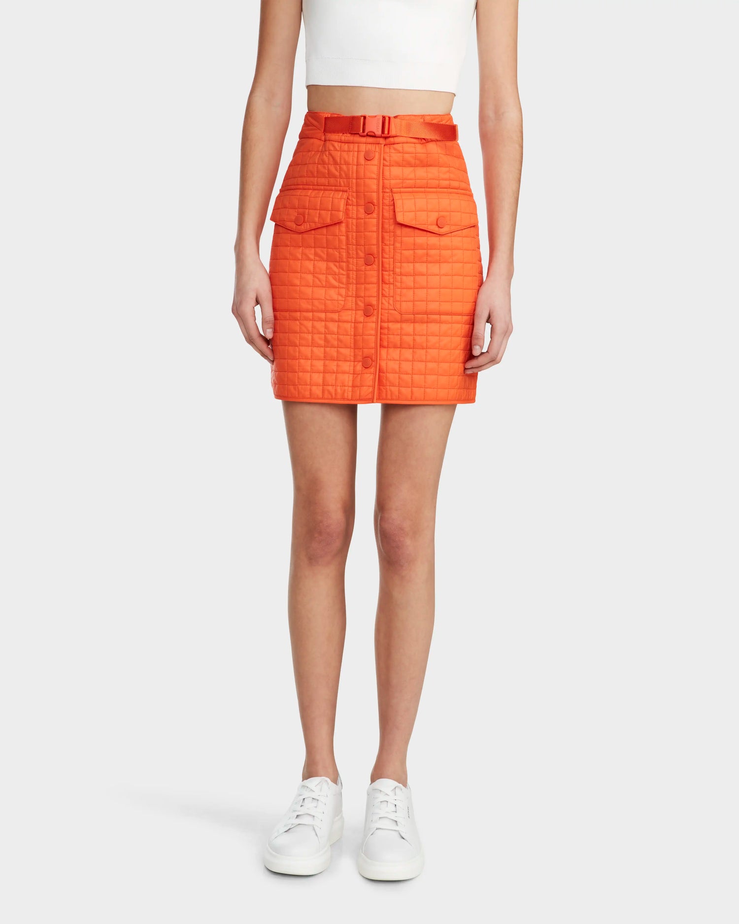 A woman wears the Rudsak SABINE - 8524063 Orange Sorbet quilted short skirt with buttons, front pockets, and a belt, paired with a white crop top and sneakers—a bold statement piece.
