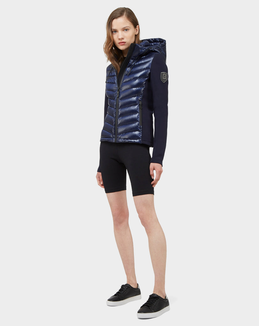 A woman faces forward wearing the Rudsak MALYA - CA EN L121006 DEEP BLUE down quilted puffer jacket with a hood, paired with black fitted shorts and sneakers. Her straight, shoulder-length hair and neutral expression complete this casual women's activewear look.