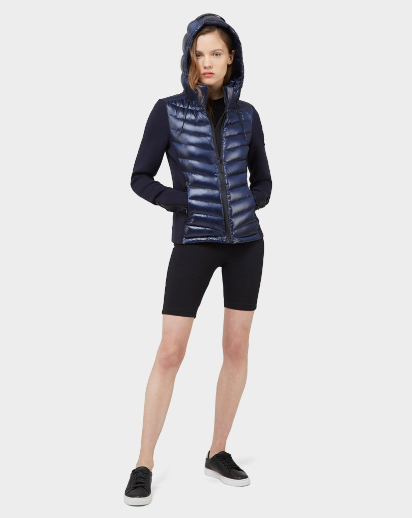 A person wears the Rudsak MALYA - CA EN L121006 DEEP BLUE, a shiny navy down quilted hooded puffer jacket, with black sleeves, cycling shorts, and sneakers—striking a modern women's activewear look against a white backdrop.