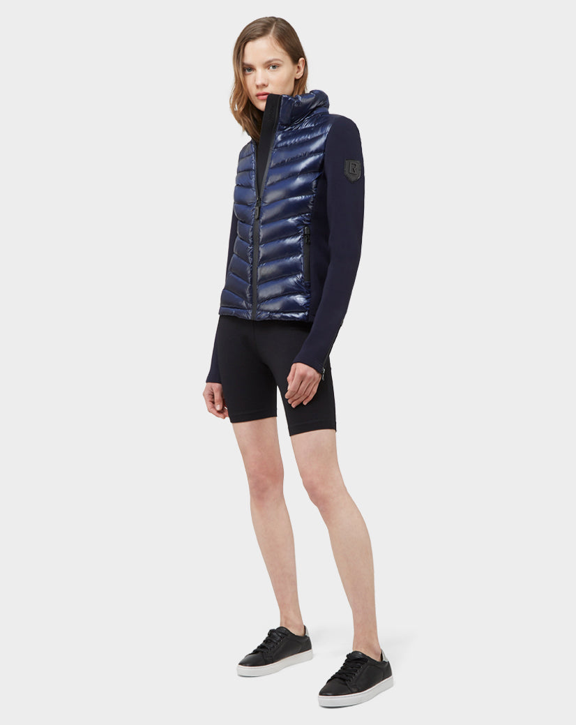 A woman with light skin and straight, shoulder-length hair faces forward against a white background wearing the Rudsak MALYA - CA EN L121006 DEEP BLUE puffer jacket, black fitted shorts, and black sneakers.