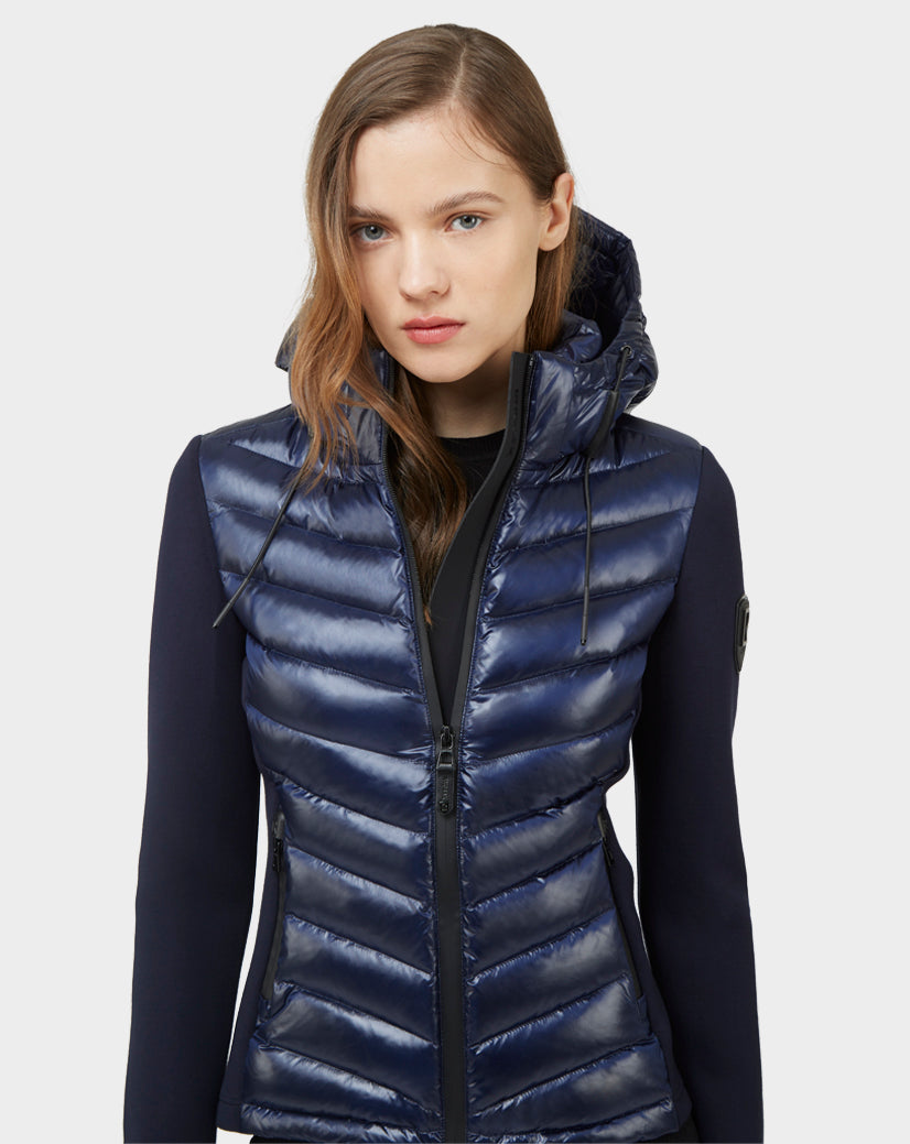 A woman with long, light brown hair models the Rudsak MALYA - CA EN L121006 DEEP BLUE down quilted puffer vest over a dark long-sleeve top, standing against a plain light gray background.
