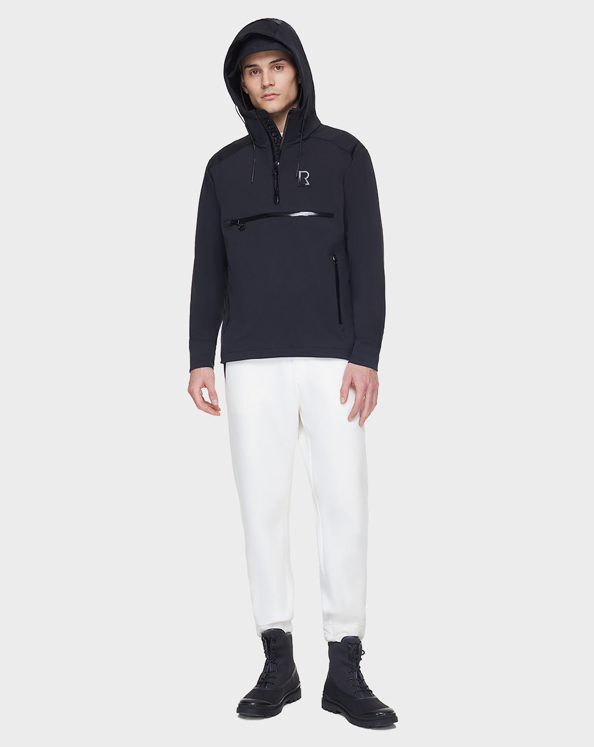 Wearing the RUDSAK AZUR - 9122001 BLACK unisex windbreaker with a front zipper pocket, paired with white pants and black boots, this look is perfect for transitional weather against a plain light background.