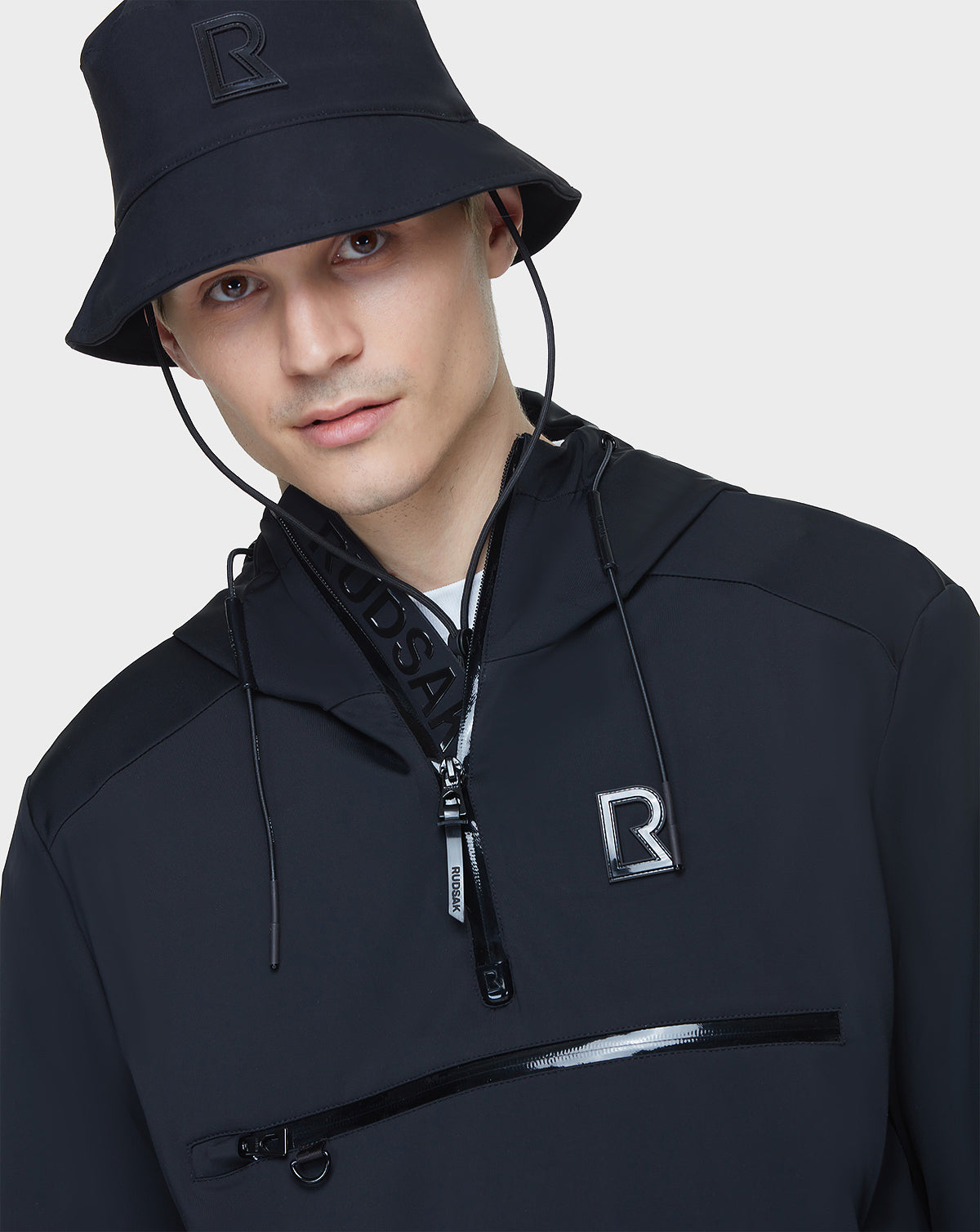 A young man in a black bucket hat and RUDSAK’s unisex AZUR - 9122001 BLACK windbreaker with R logos, a half-zip front, and zippered pocket stands against a plain white background—perfect for transitional weather.