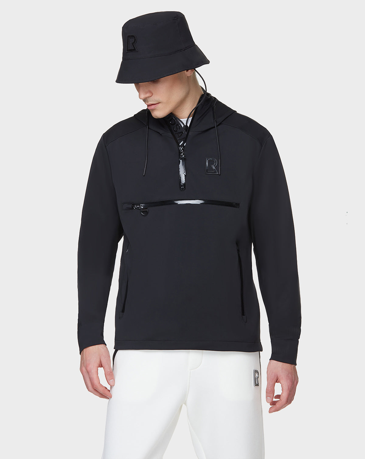 Dressed in the RUDSAK AZUR - 9122001 BLACK windbreaker bucket hat and matching hooded pullover with front zipper pocket, plus white pants, the look is finished with subtle R logos—a staple for transitional weather.