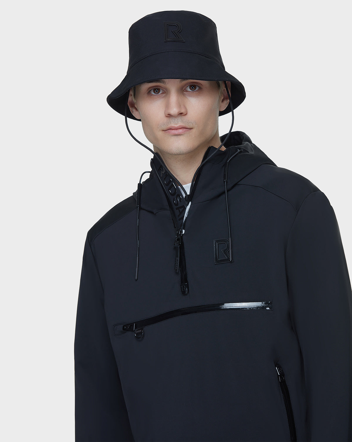 A person wears the AZUR - 9122001 BLACK unisex windbreaker jacket and a matching black bucket hat by RUDSAK, both with a subtle R logo, standing against a plain white background—ideal for transitional weather.