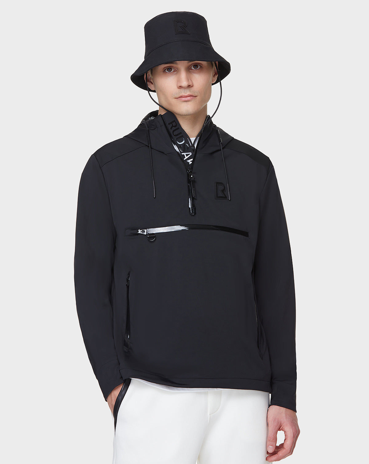A young man wearing the RUDSAK AZUR - 9122001 BLACK unisex windbreaker bucket hat and zip-up jacket with logo details, paired with white pants, stands against a plain light gray background.