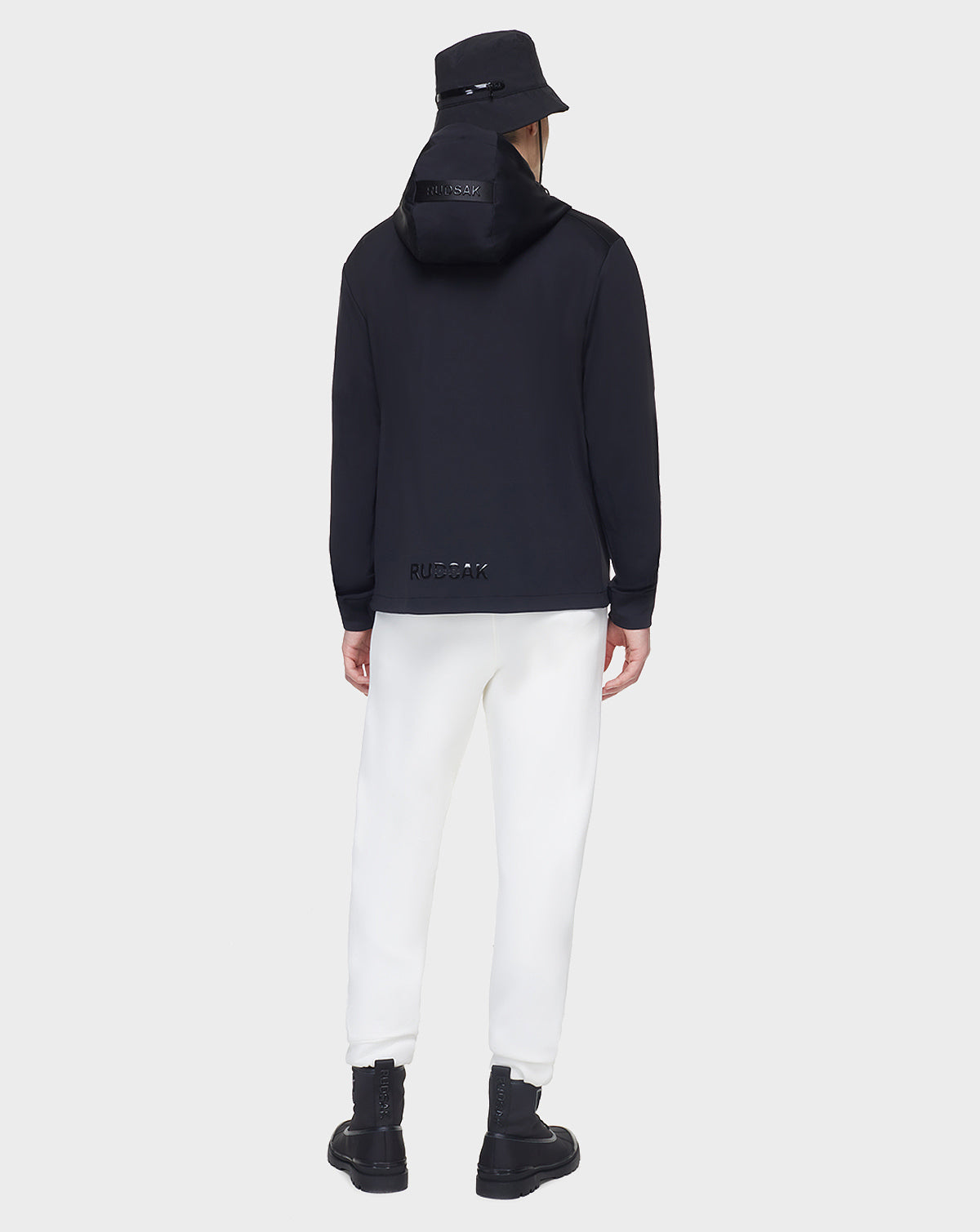 Wearing the RUDSAK AZUR - 9122001 BLACK bucket hat, a person faces away in a unisex windbreaker, white pants, and black boots against a plain white background—a perfect transitional weather staple.