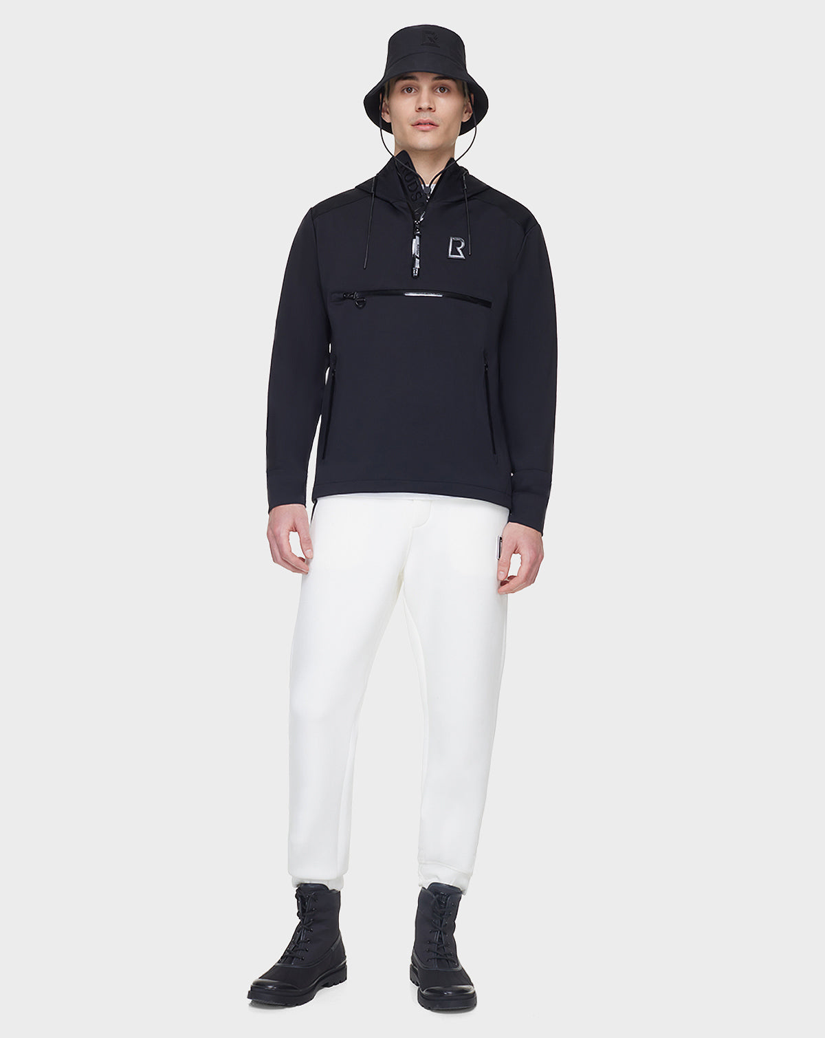 A person stands against a plain background wearing the RUDSAK AZUR - 9122001 BLACK unisex windbreaker pullover with a zippered chest pocket, paired with a black bucket hat, white pants, and black boots.
