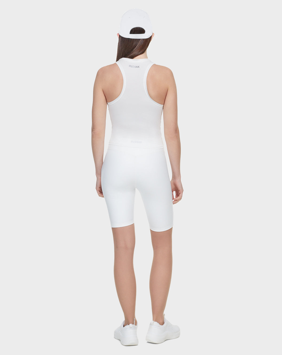 A woman faces away in Rudsak's SABIE - 8522020 WHITE: a white cap, sleeveless top, bicycle shorts, and sneakers, set against a plain light background.