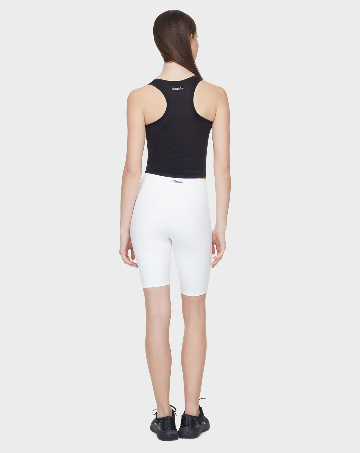 A woman with straight brown hair, wearing the Rudsak SABIE - 8522020 BLACK sleeveless tank top stands facing away against a plain light gray background.