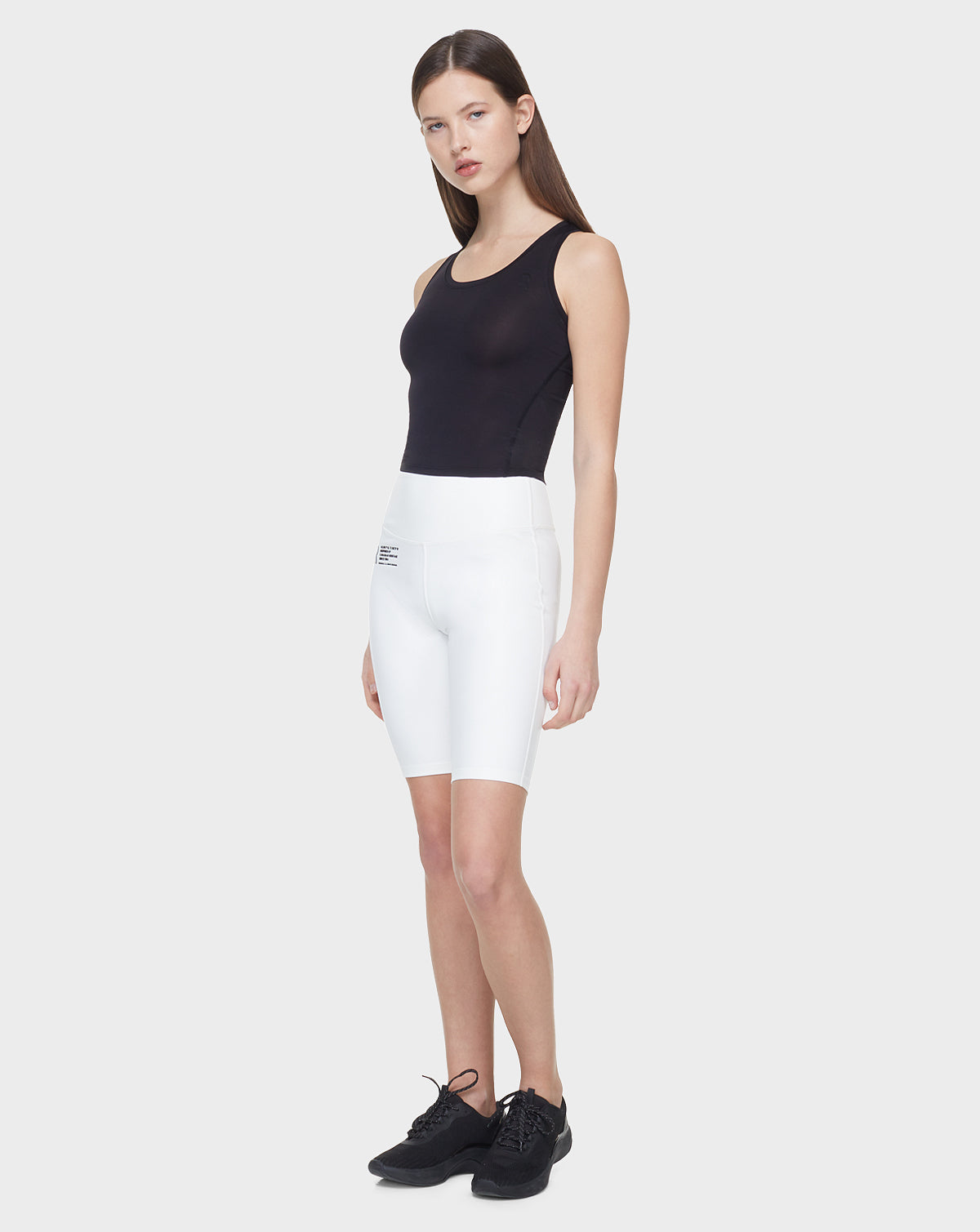 A woman with straight brown hair models the Rudsak SABIE - 8522020 BLACK sleeveless top, paired with white high-waisted biker shorts and black sneakers, standing against a plain light gray background.