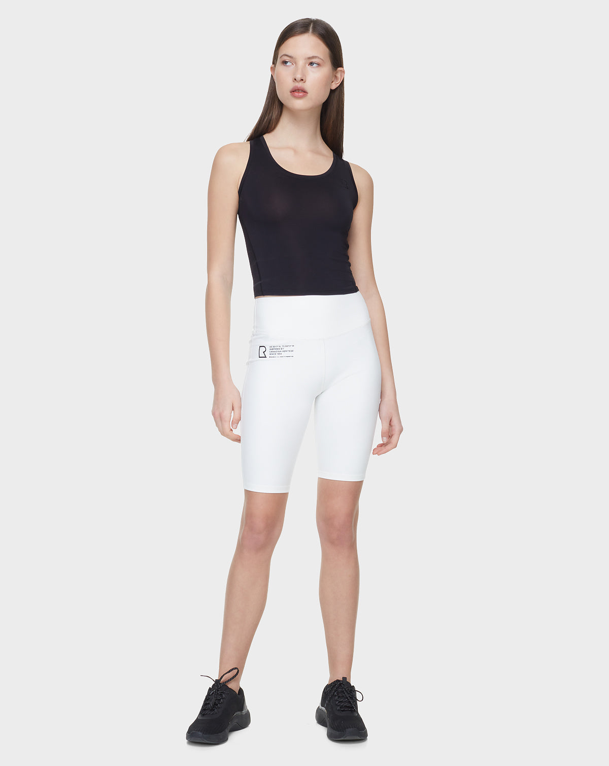 A woman with straight brown hair looks slightly left, wearing the Rudsak SABIE - 8522020 BLACK sleeveless tank top against a plain background. She pairs it with white high-waisted bike shorts and black sneakers.