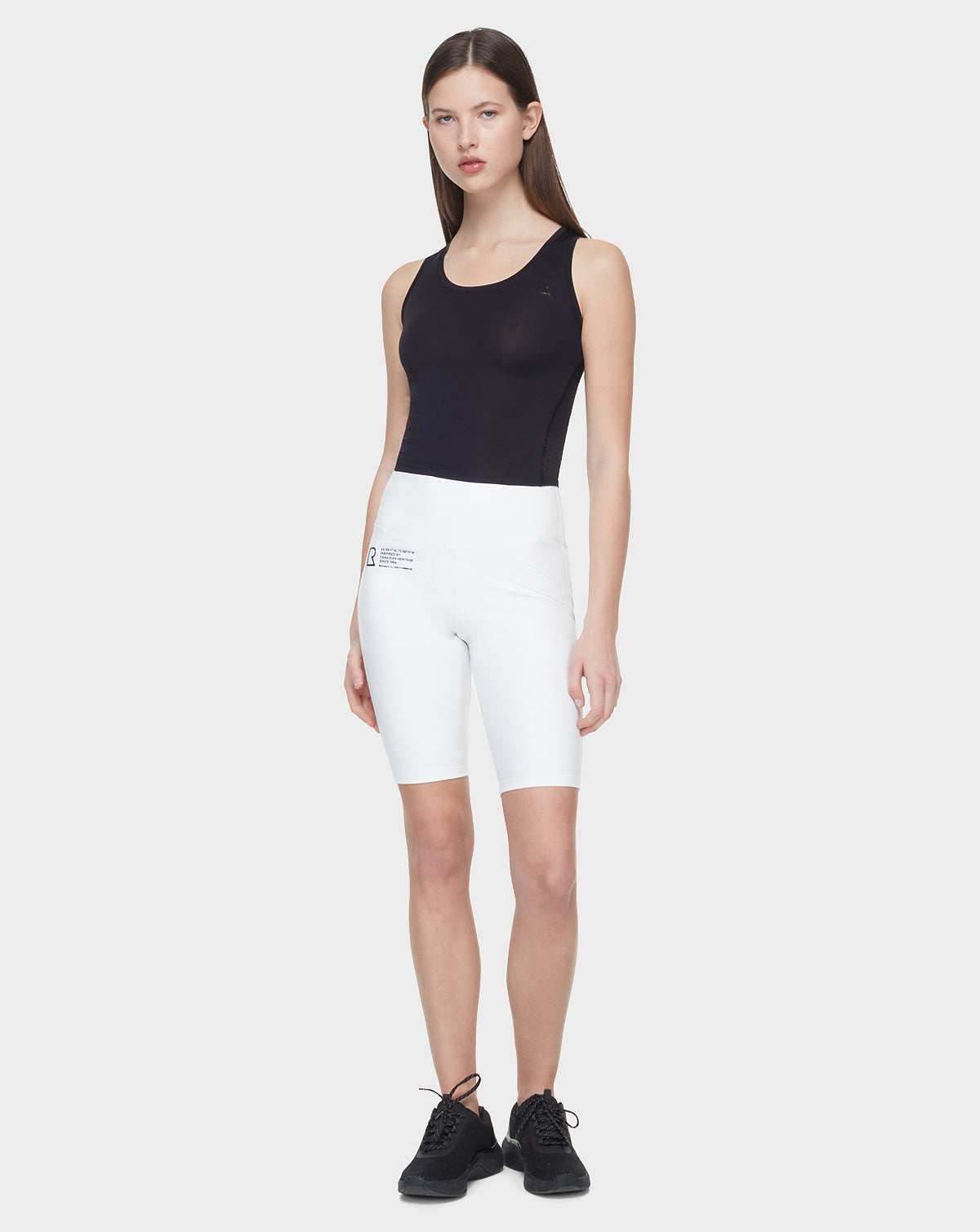 A woman with straight brown hair wears Rudsak's SABIE - 8522020 BLACK sleeveless top, white high-waisted cycling shorts with a logo on the left thigh, and black sneakers, standing against a plain light gray background.