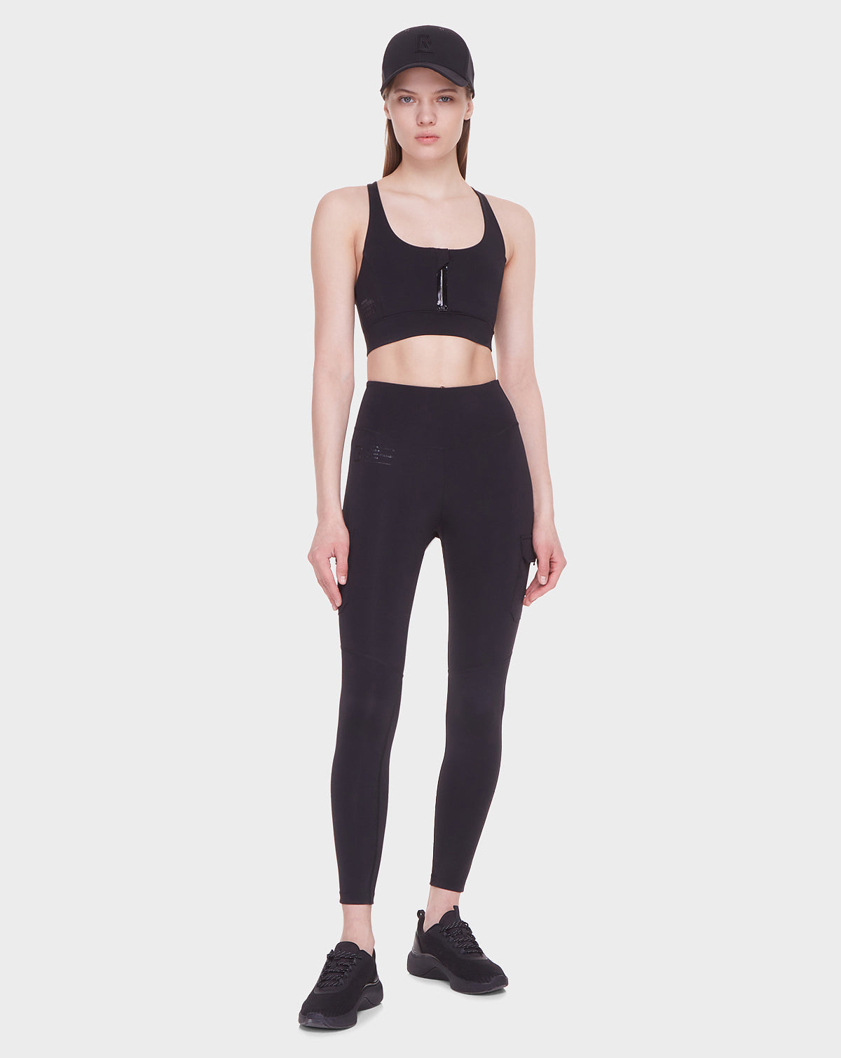 A woman models Rudsak’s SIEN - 8521001 BLACK against a plain background, wearing the black activewear set with a neutral expression and her arms at her sides.