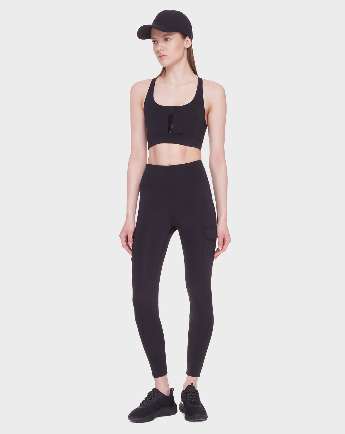 A woman wearing the SIEN - 8521001 BLACK sports bra by Rudsak, paired with high-rise stretch leggings, black sneakers, and a black baseball cap stands against a plain white background.