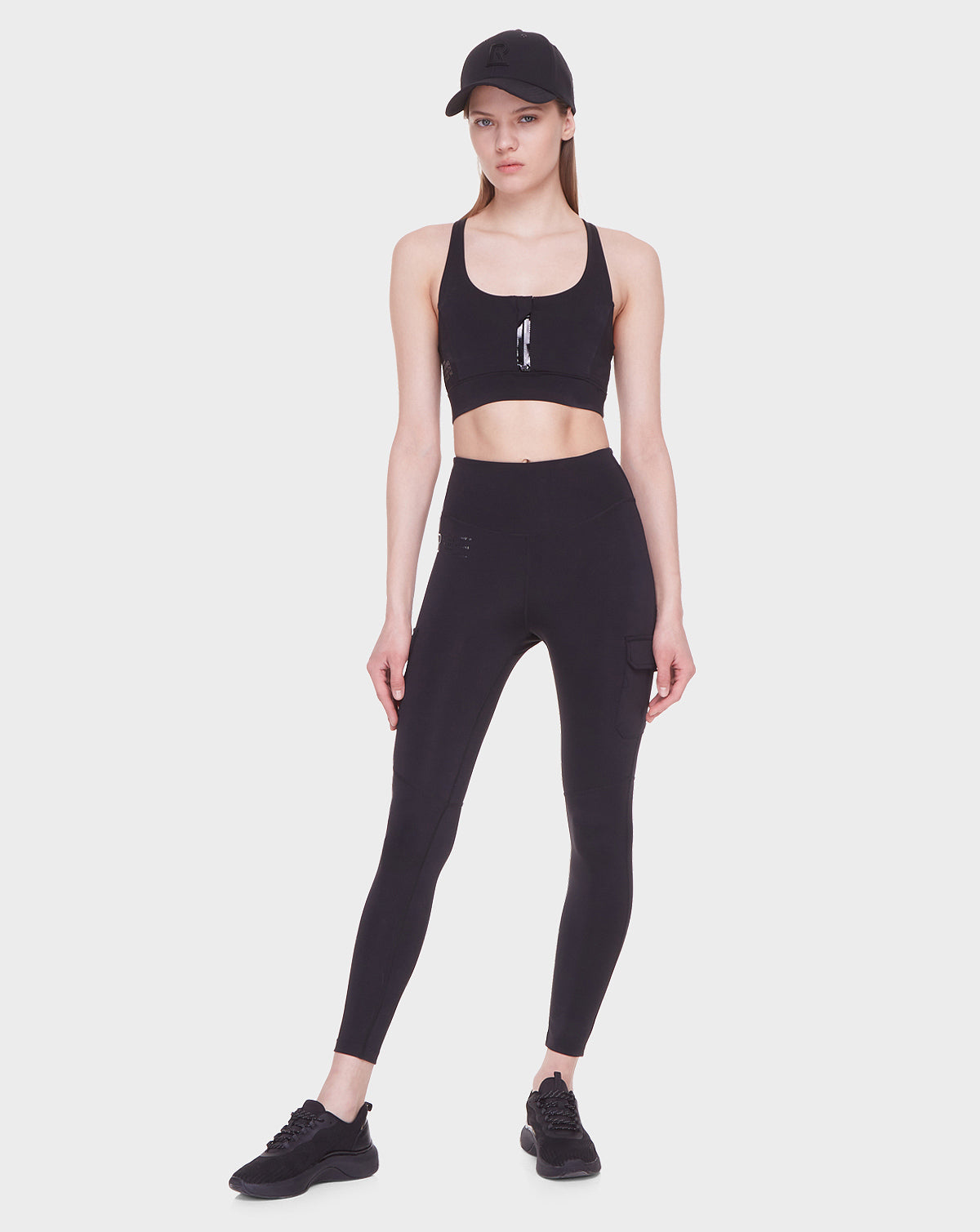 A woman wears the Rudsak SIEN - 8521001 BLACK sports bra and high-rise leggings, paired with black sneakers and a cap, standing against a white background, showcasing sleek modern activewear with a composed look.