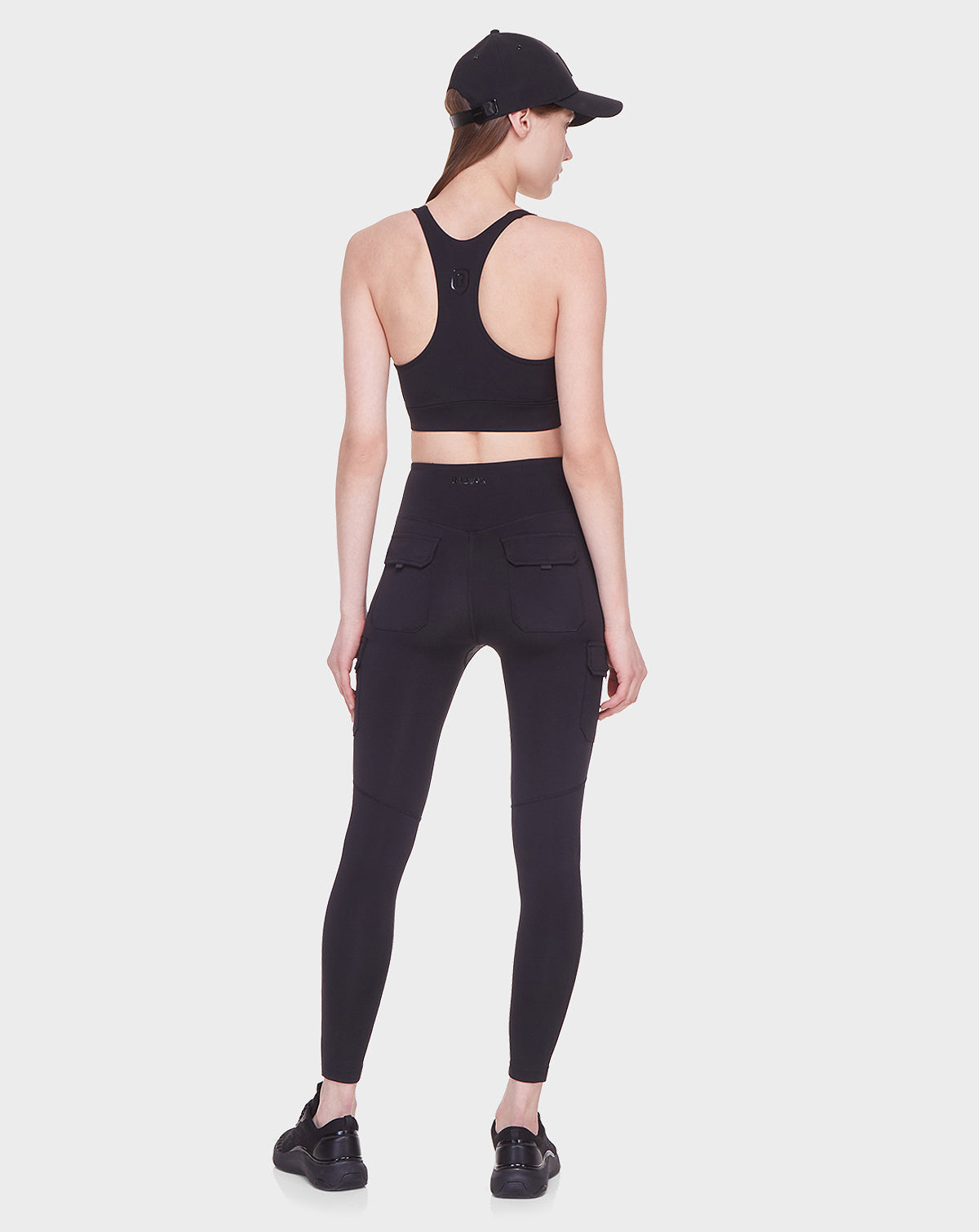 A woman, facing away, models Rudsak’s SIEN - 8521001 BLACK: a black sports bra and high-rise leggings with side pockets. She finishes the activewear look with black sneakers and a cap, all set on a plain white background.