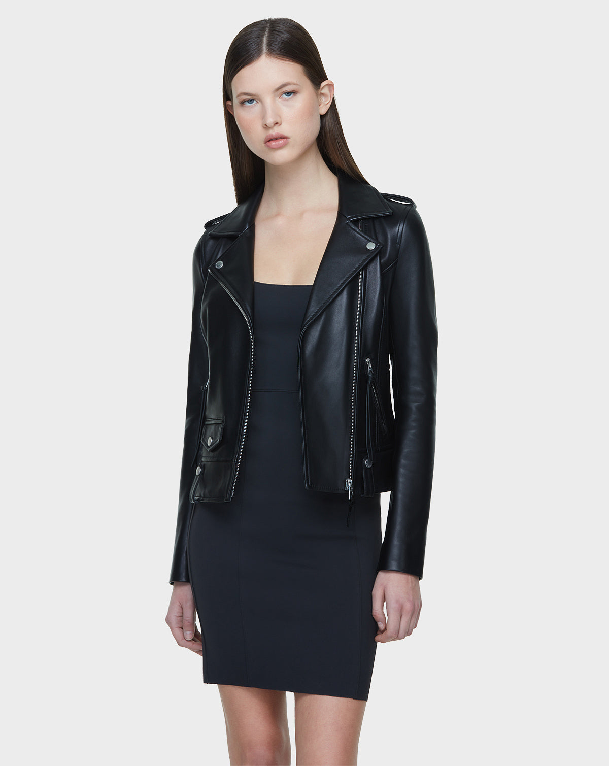 A woman with straight brown hair faces forward in a fitted MERGO - EN US Black 8118525 dress by Rudsak International, layered under a black biker jacket with an asymmetrical zip, set against a plain light gray background.