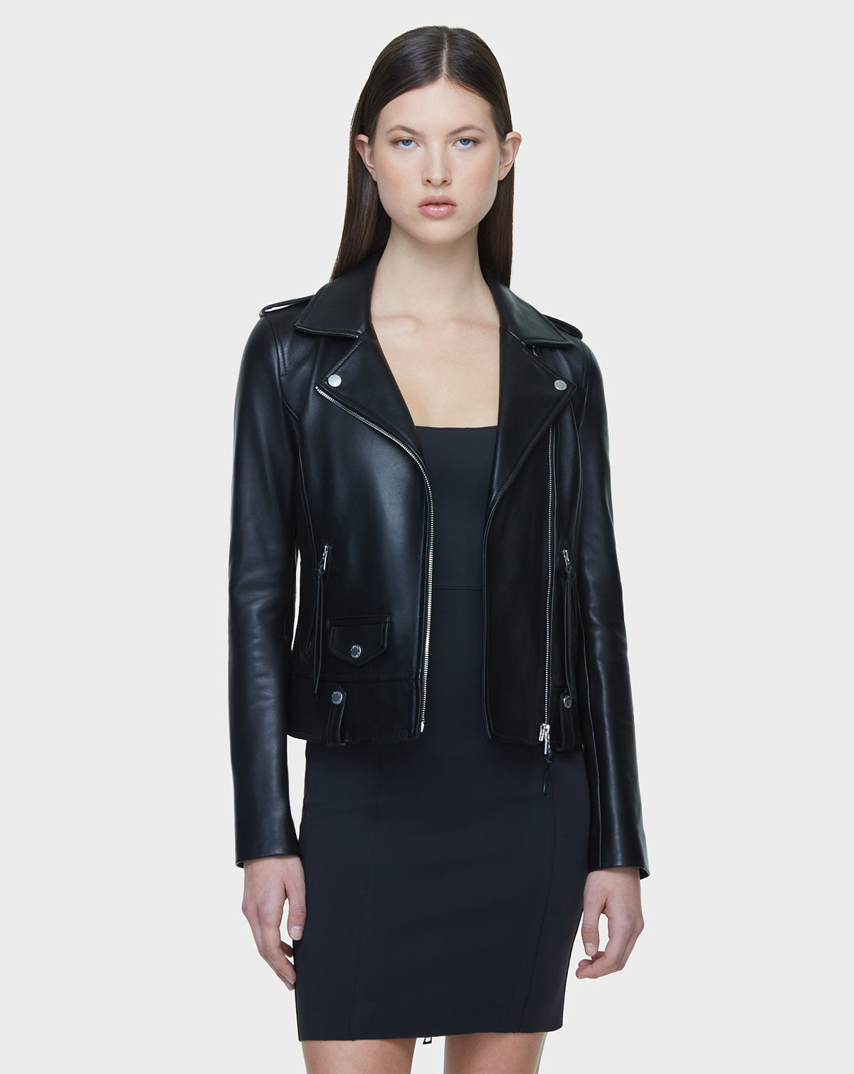 A woman with straight brown hair is wearing the MERGO – EN US Black 8118525 biker jacket by Rudsak International over a black dress, standing against a plain light gray background.