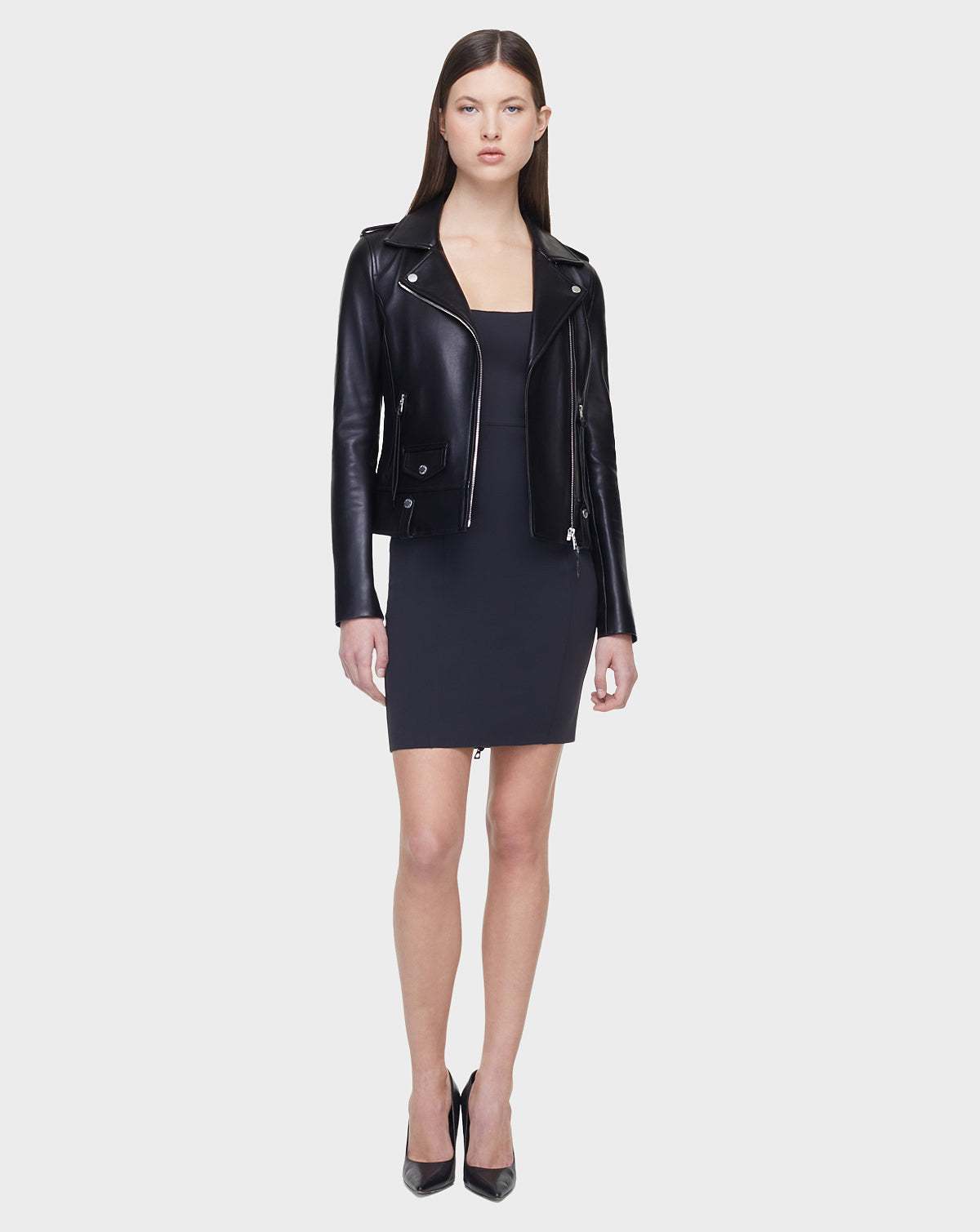 A woman faces forward, wearing the MERGO - EN US Black 8118525 biker jacket by Rudsak International over a fitted dark dress and black high heels, standing against a plain light gray background.