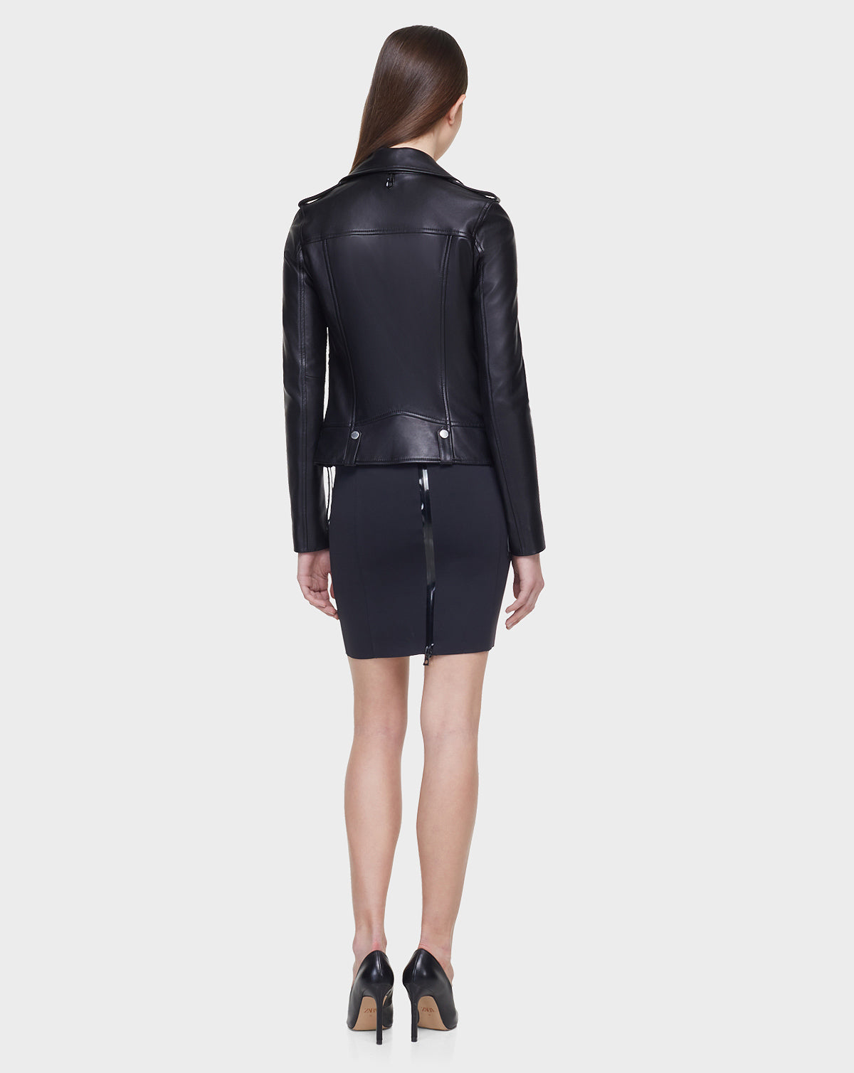 A woman with long brown hair faces away, wearing the MERGO - EN US Black 8118525 biker jacket and skirt by Rudsak International with a visible back zipper. She completes the look with black high heels, set against a plain white background.