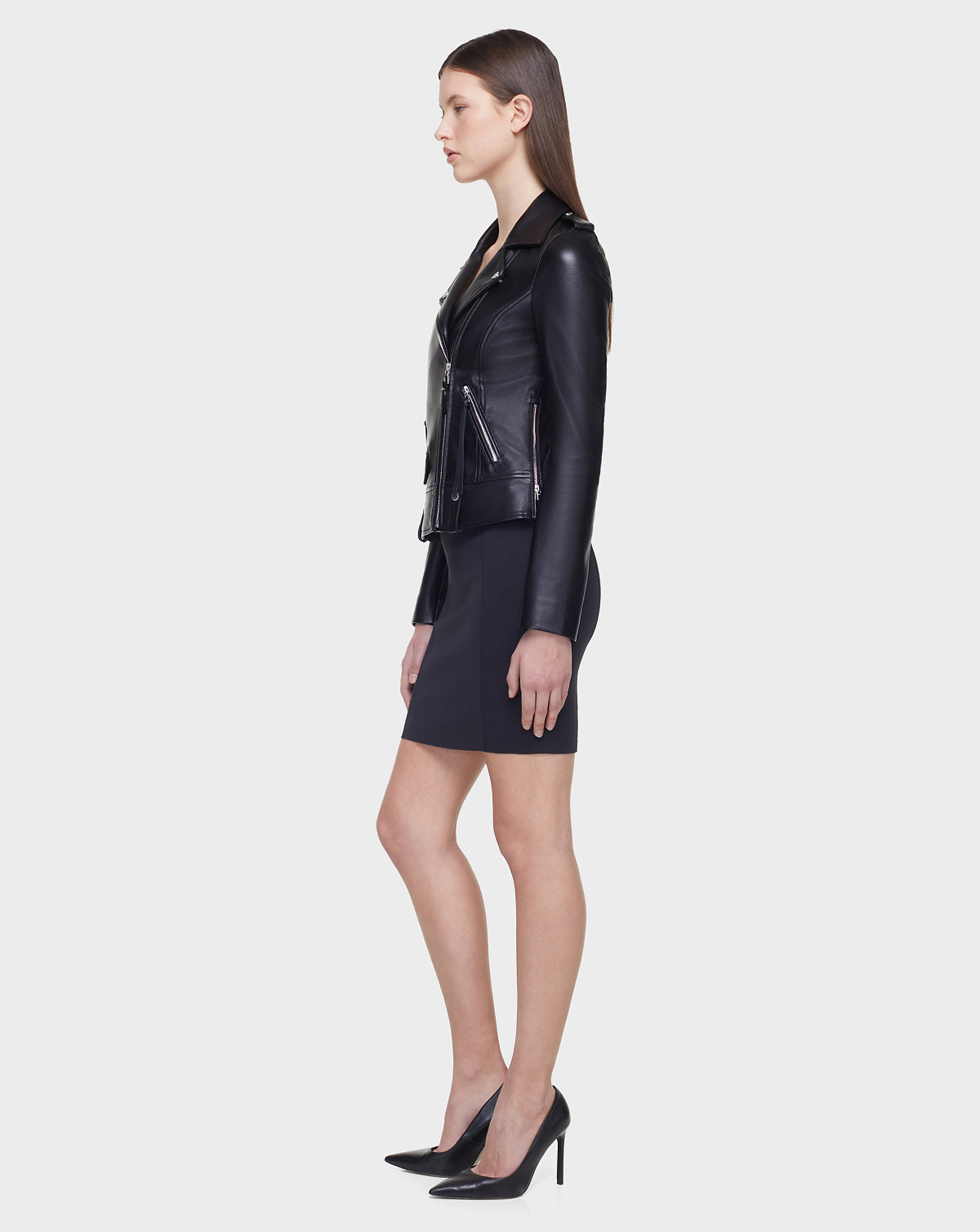 A woman with straight hair, facing left in profile against a plain background, wears the MERGO - EN US Black 8118525 biker jacket by Rudsak International with a dark skirt and black high-heeled shoes.