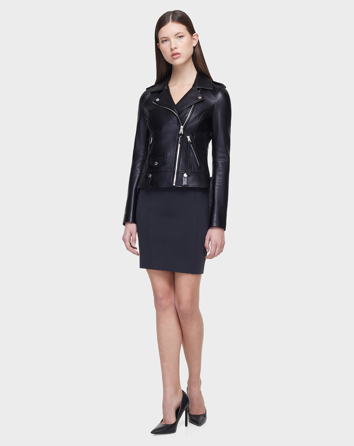 A woman faces forward wearing the MERGO - EN US Black 8118525 leather biker jacket by Rudsak International, styled with a black skirt and high-heeled shoes, set against a plain light gray background.